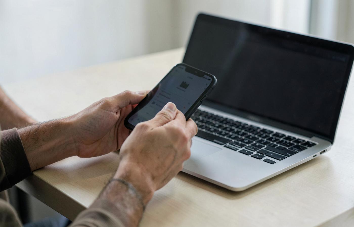 Hands holding a smartphone beside an open laptop on a desk
