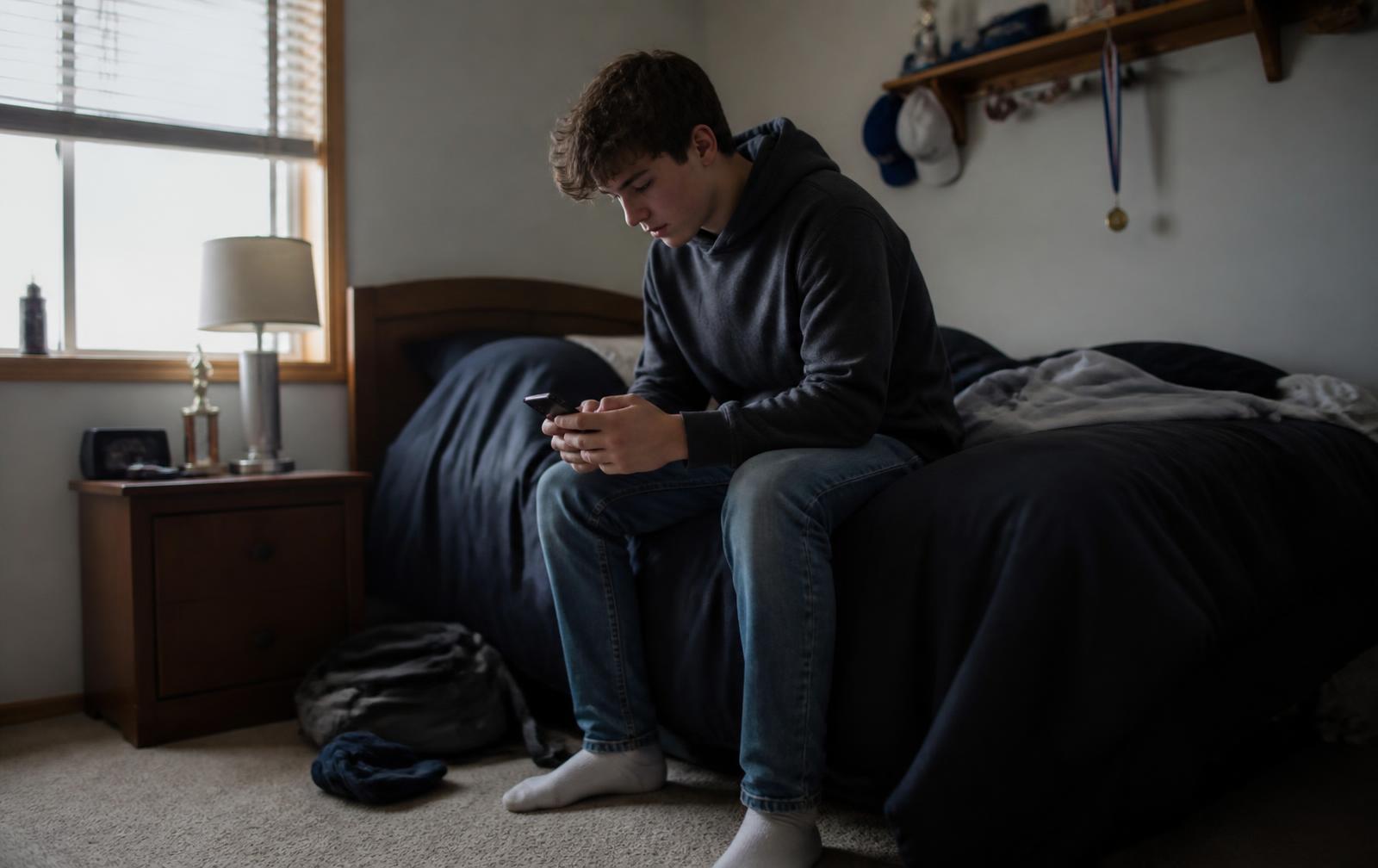 A teenager sitting on the edge of a bed in a suburban bedroom, looking at his phone